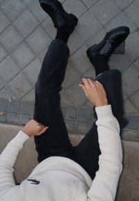 Μαύρα μποτάκια μέχρι τον αστράγαλο, σκούρο τζιν και ένα ριγέ κρεμ πουλόβερ με λογότυπο. Η σκηνή περιλαμβάνει ένα πλακόστρωτο έδαφος με μερικά φύλλα. Χέρι resting σε ένα πόδι.