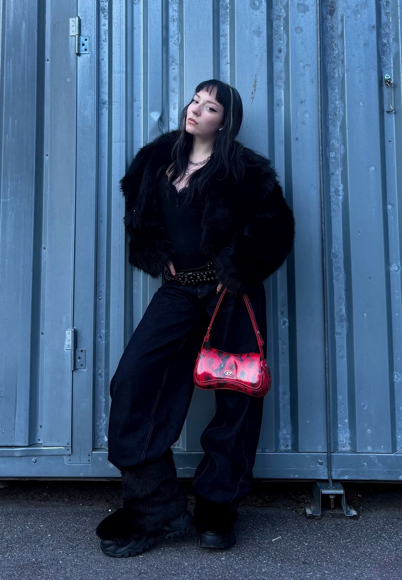 Veste en fausse fourrure noire, haut noir, pantalon en denim à jambes larges, bottes à plateforme noires et sac à main rouge à motif. Arrière-plan : conteneur métallique gris.