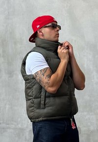 Gilet imbottito verde con colletto alto, indossato sopra una camicia bianca. Caratterizzato da una zip, tasche laterali e un portachiavi decorativo. Cappellino da baseball rosso.