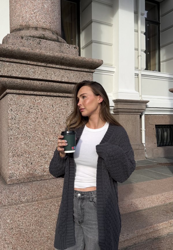 Grå strikket cardigan med overdrevne ærmer, båret over en hvid top, holdt med en take-away kaffekop foran en baggrund af en stenspille.