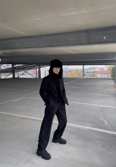 Traje negro a medida con un suéter de cuello alto negro, capucha grande y peluda, y gafas de sol reflectantes. Zapatos negros con suela gruesa. Fondo de estructura de estacionamiento urbana.