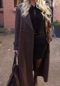 Cappotto lungo in camoscio marrone con bottoni doppiopetto, abbinato a un top corto nero e una minigonna. Tiene una borsa abbinata. Scarpe basse.
