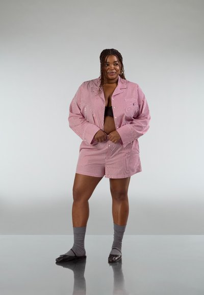 Conjunto de pijama a rayas rojas y blancas con camisa abotonada y pantalones cortos. El modelo lleva calcetas grises y zapatillas negras, de pie frente a un fondo neutro.