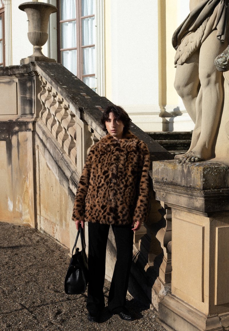 Personne portant un manteau en fourrure imprimé léopard marron et un pantalon noir, tenant un sac noir, debout à côté de marches en pierre et des jambes d'une statue.