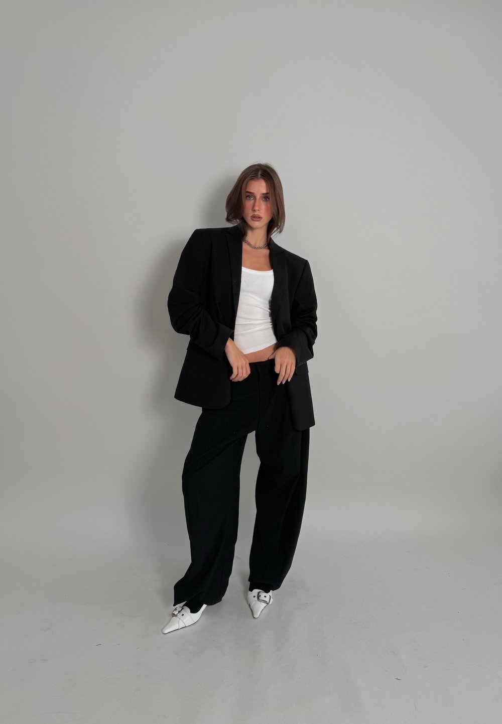 Blazer noir sur un débardeur blanc ajusté, associé à un pantalon noir ample et des chaussures blanches à bout pointu. Tissu lisse et coupe surdimensionnée.