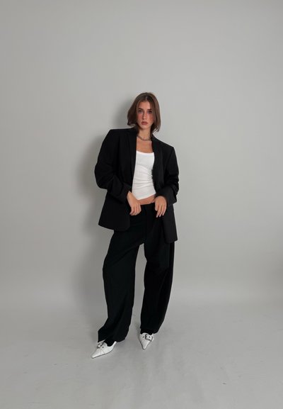 Blazer negro sobre una camiseta sin mangas blanca ajustada, combinada con pantalones negros holgados y zapatos blancos puntiagudos. Tela lisa y ajuste holgado.