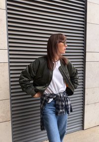 Veste bomber vert olive à finition lisse, dotée d'une fermeture éclair, de poches latérales et de poignets côtelés, portée sur un T-shirt blanc et une chemise à carreaux nouée.