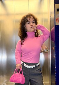 Ružičasti rolka s dugim rukavima, sive hlače visokog struka, bijeli remen, držeći malu ružičastu torbicu, u srebrnom dizalu.