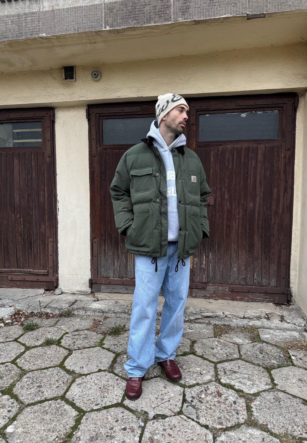 Doudoune verte avec poches avant, sweat à capuche gris, jean bleu ample et chaussures bordeaux, debout devant des portes en bois et un trottoir.