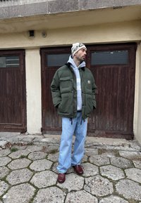 Doudoune verte avec poches avant, sweat à capuche gris, jean bleu ample et chaussures bordeaux, debout devant des portes en bois et un trottoir.