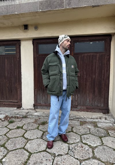 Chaqueta acolchada verde con bolsillos delanteros, sudadera con capucha gris, vaqueros azules holgados y zapatos burdeos, de pie frente a puertas de madera y pavimento.