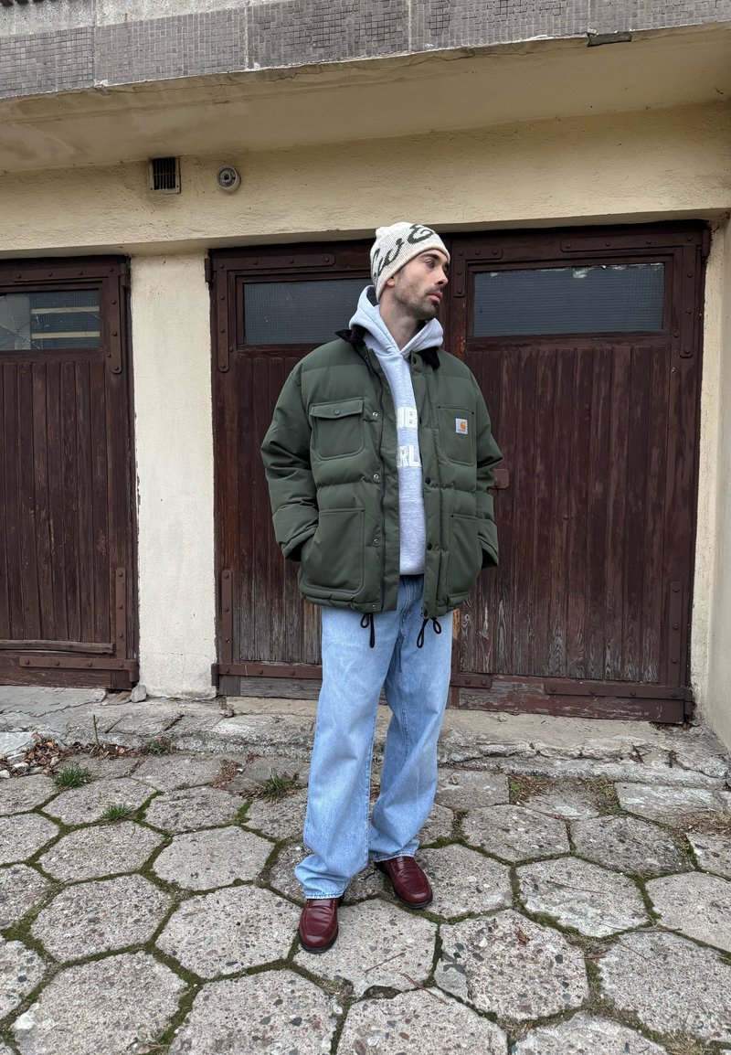 Doudoune verte avec poches avant, sweat à capuche gris, jean bleu ample et chaussures bordeaux, debout devant des portes en bois et un trottoir.