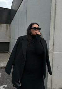 Črn oversize blazer preko črne rebraste turtleneck majice, v kombinaciji s črnimi sončnimi naočniki. Gladke teksture, minimalističen dizajn, brez vidnih vzorcev.
