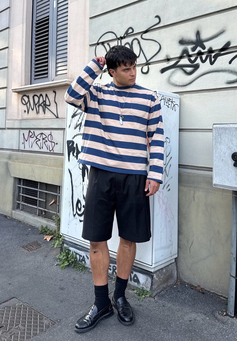 Camicia a maniche lunghe a righe crema e blu, abbinata a pantaloni corti neri. Gli accessori includono un ciondolo d'argento e mocassini neri con calzini.