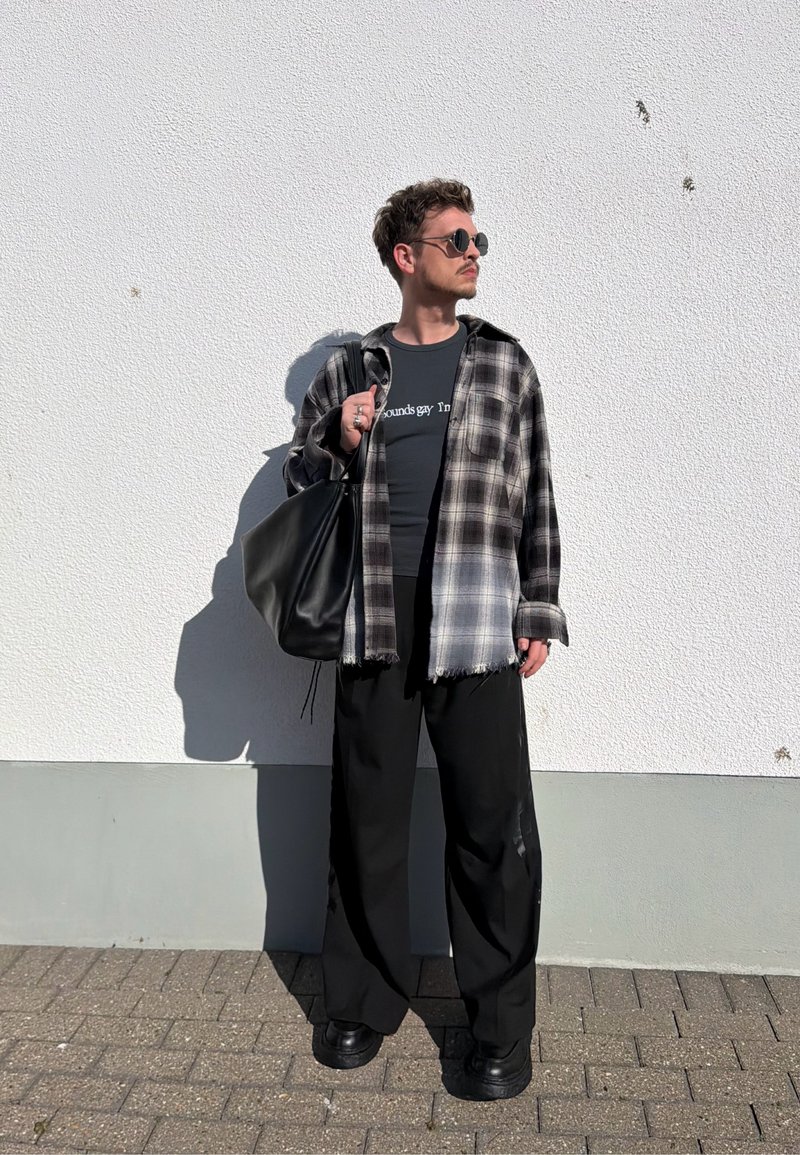 Homme portant des lunettes de soleil rondes, chemise à carreaux, pantalon large noir, tenant un sac noir, debout contre un mur blanc texturé au soleil.