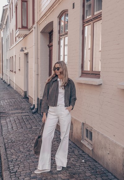 Mujer con gafas de sol, chaqueta oscura, pantalones blancos y zapatillas parada en una calle empedrada junto a un edificio de ladrillos beige con ventanas.