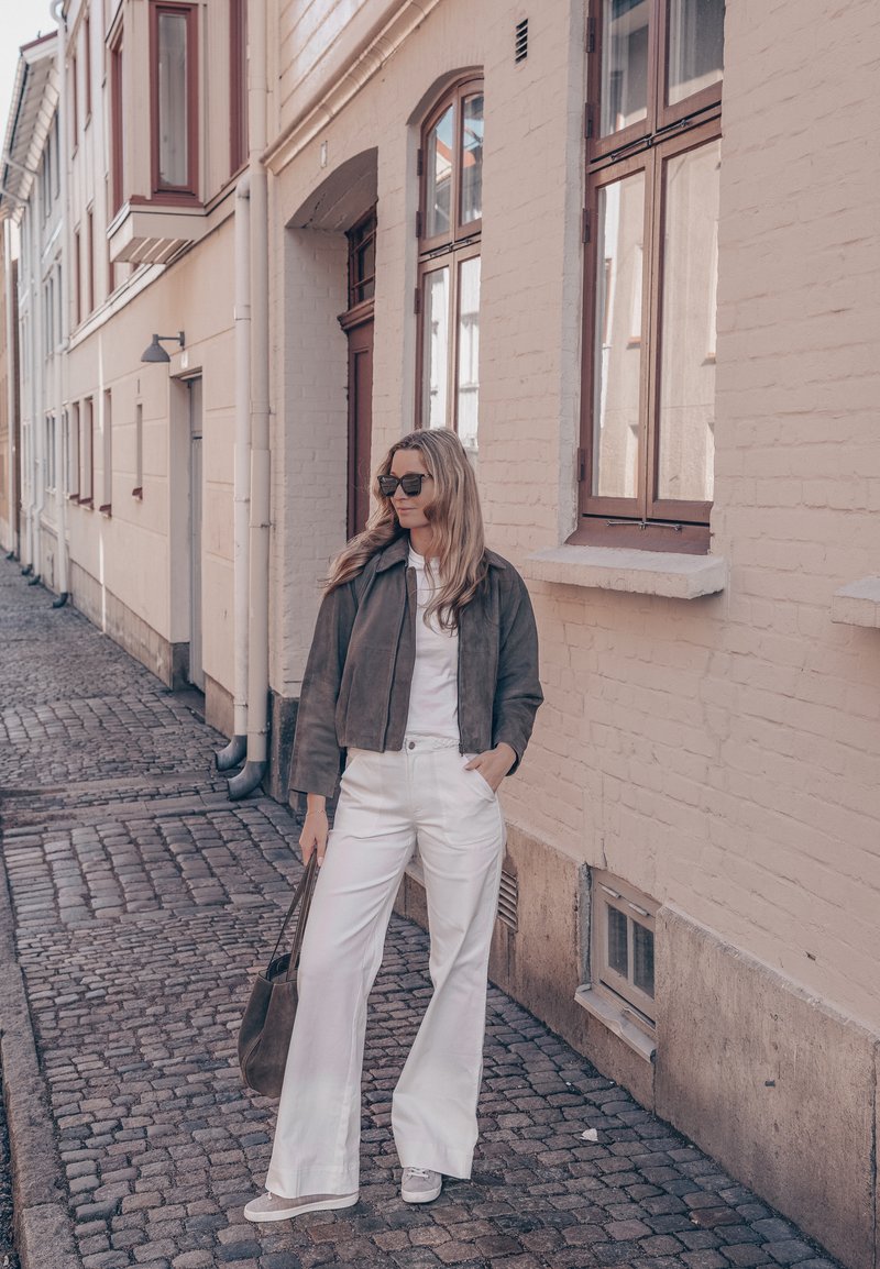 Femme portant des lunettes de soleil, une veste sombre, un pantalon blanc et des baskets, se tenant sur une rue pavée à côté d'un bâtiment en brique beige avec des fenêtres.