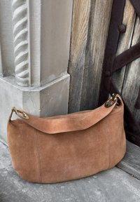 Helle Wildleder-Umhängetasche mit geschwungenem Design, ausgestattet mit einem Reißverschluss und einem breiten Tragegurt mit goldfarbenen Details. Strukturierte Oberfläche.