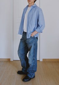 Blåvit randig skjorta över en vit t-shirt, i kombination med lösa blå denimjeans och svarta skor. Stående på ett trägolv.