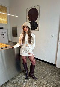 Femme portant une tenue crème, des collants à motifs, des bottes bordeaux et un chapeau seau, appuyée sur un comptoir en métal avec des produits de boulangerie dans un décor intérieur minimaliste.