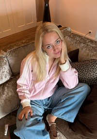 Veste zippée rose avec des rayures blanches, associée à un pantalon en denim à jambes larges. Assise sur un canapé moelleux avec des coussins à imprimé léopard. Chaussons marron.