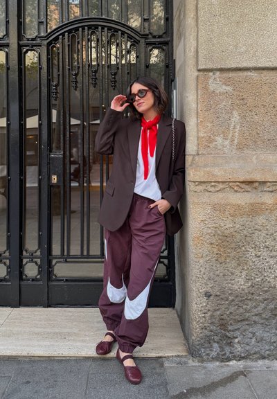 Mujer con blazer oscuro, bufanda roja, pantalones burdeos y blancos, y gafas de sol apoyada contra una pared de piedra junto a una entrada con verja negra.