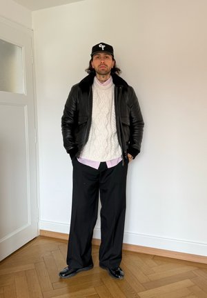 Hombre de pie en interiores vistiendo una gorra negra, chaqueta de cuero negra con cuello de piel, suéter blanco de punto con ochos, camisa rosa, pantalones negros y zapatos negros.