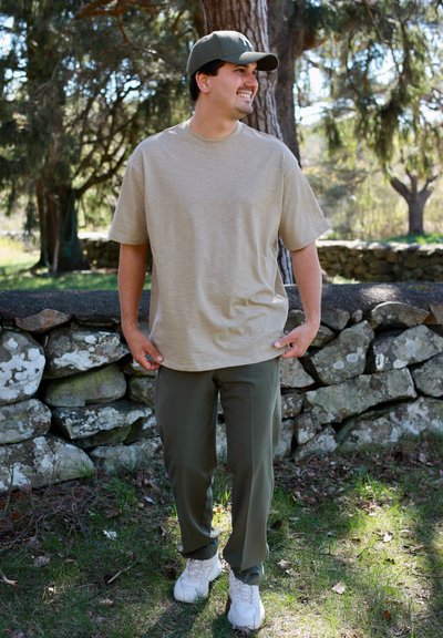 Hombre con camiseta beige, pantalones verde oliva y gorra verde, de pie al aire libre cerca de una pared de piedra con árboles al fondo.