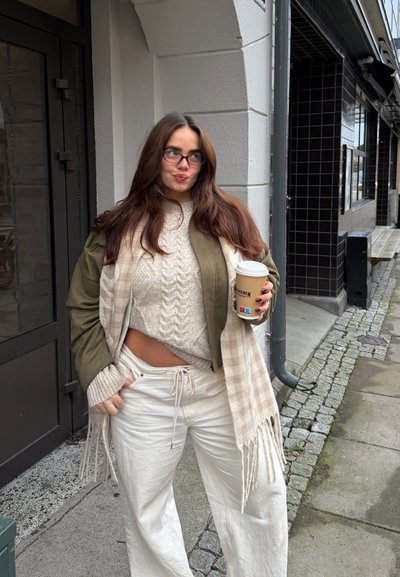 Una mujer con una chaqueta verde oliva, suéter de punto crema y pantalones blancos sostiene una taza de café. Lleva una bufanda de cuadros con flecos.