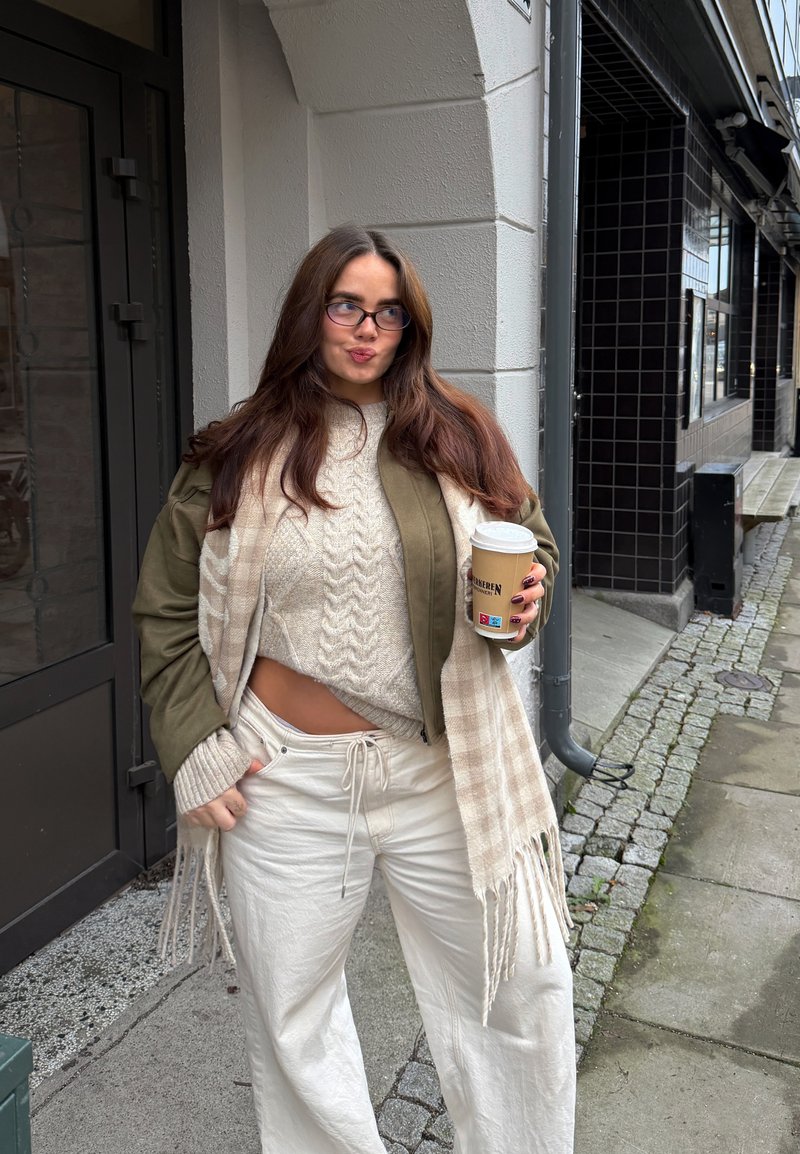 En kvinde iført en olivengrøn jakke, en cremefarvet strikket sweater og hvide bukser holder en kaffekop. Hun har et ternet tørklæde med frynser.