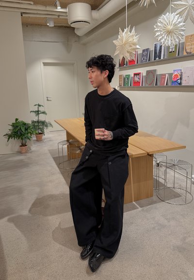 Camisa negra de manga larga con bolsillo, combinada con pantalones anchos negros con detalles de botones. Ambiente minimalista con mesa de madera y plantas.