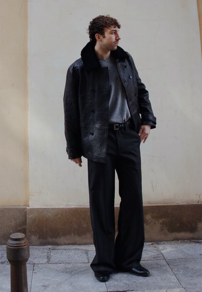 Hombre con cabello rizado que lleva una chaqueta de cuero negra con cuello de piel, suéter gris, pantalones anchos negros y zapatos negros, de pie sobre el pavimento.