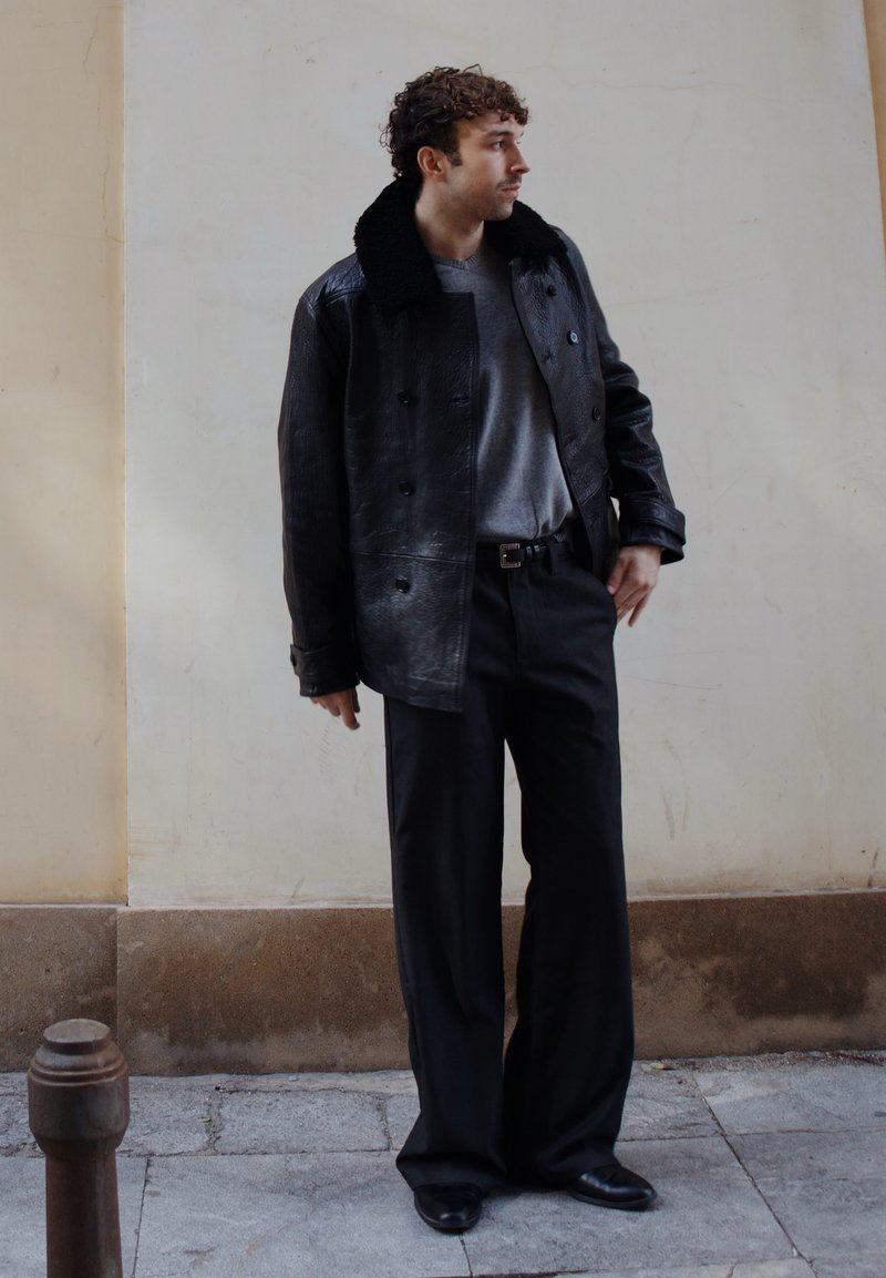 Homme aux cheveux bouclés portant une veste en cuir noire avec col en fourrure, un pull gris, un pantalon large noir et des chaussures noires, debout sur le trottoir.
