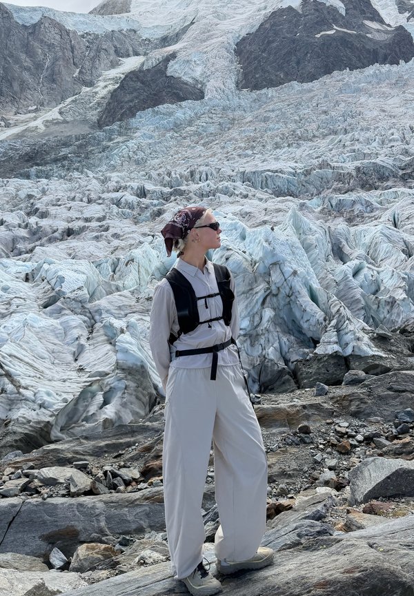 Person som bär lätt vandringskläder, solglasögon och bandana står på stenig mark med en stor glaciär och berg i bakgrunden.