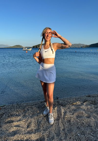 Sujetador deportivo blanco con logo negro de Nike, falda corta blanca a juego y zapatillas blancas. Posado junto al agua con un cielo azul claro de fondo.