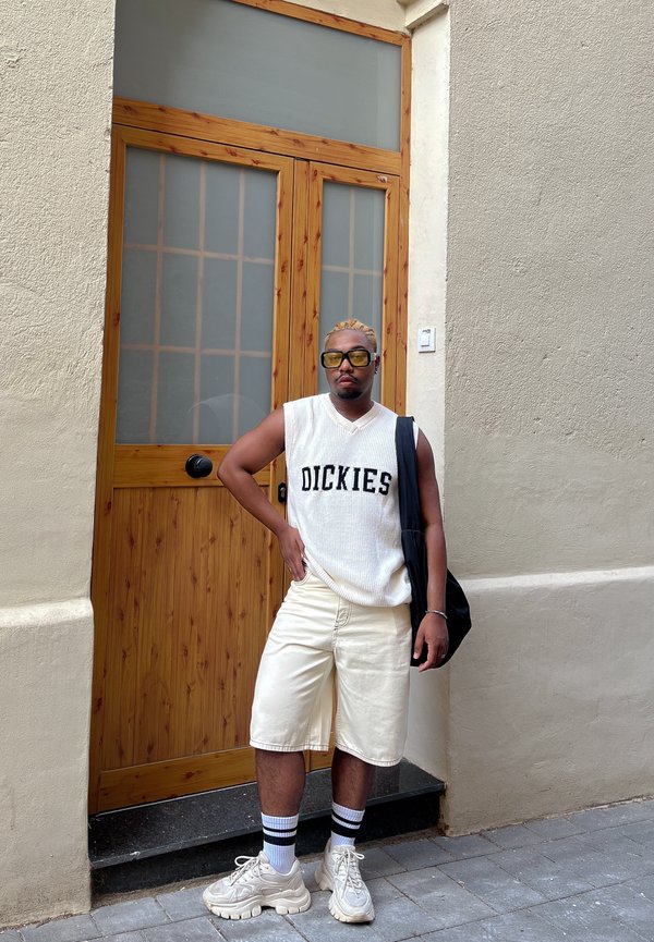 Top de punto sin mangas blanco con texto "DICKIES", pantalones cortos crema y zapatillas claras. Combinado con un bolso oscuro y calcetas a rayas, de pie junto a una puerta de madera.