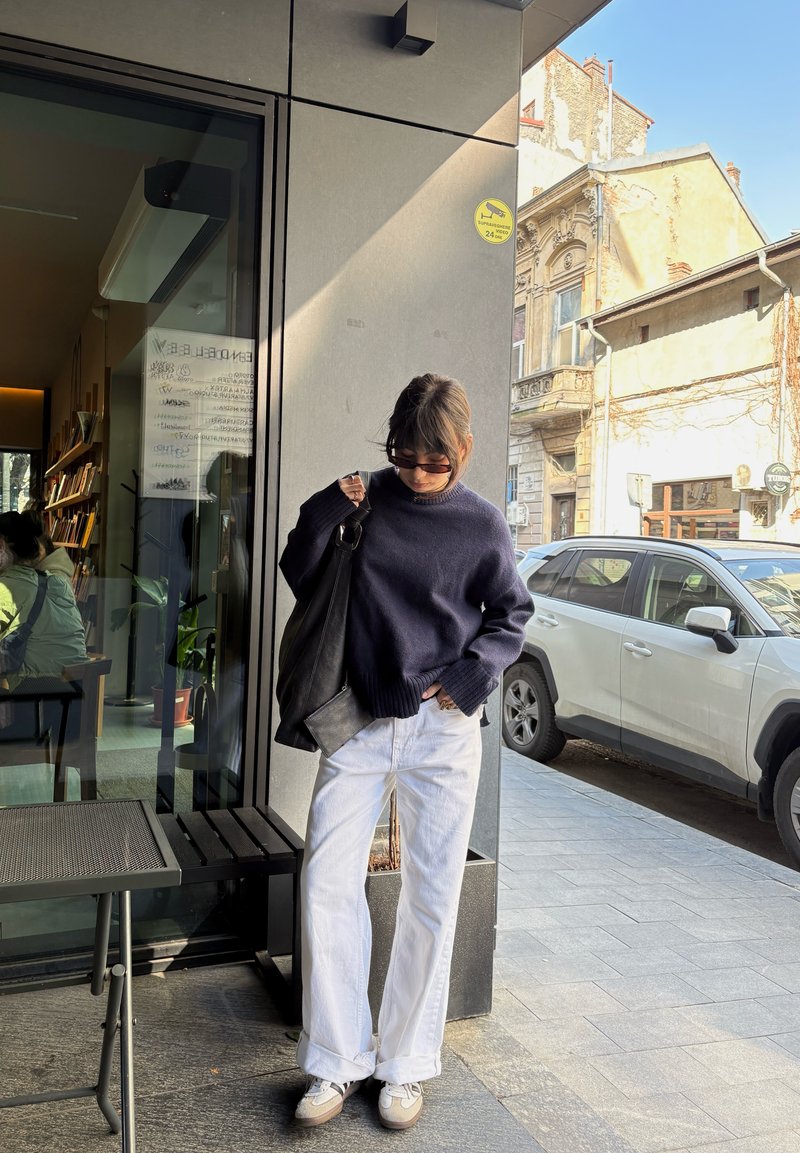 Personne portant des lunettes de soleil, un pull bleu marine, un pantalon blanc et des baskets, debout près de l'entrée d'un café avec une voiture blanche garée et de vieux bâtiments en arrière-plan.