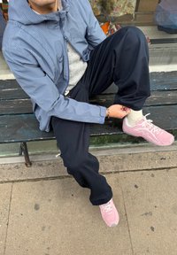 Veste à carreaux bleu clair zippée par-dessus un pull gris, pantalon foncé et baskets roses, assis sur un banc en bois en plein air.