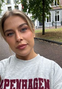 Sudadera gris con lettering rojo "COPENHAGUE", el fondo incluye árboles y edificios, luz natural, primer plano de la cara.