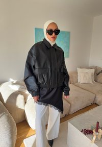 Veste en cuir noire courte avec poignets élastiques, portée sur un haut en satin noir bordé de dentelle, associée à un pantalon blanc à jambes larges et des lunettes de soleil.