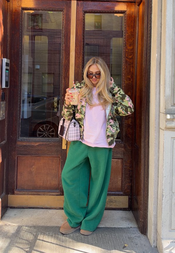 Une femme portant des lunettes de soleil se tient dans une embrasure de porte en bois, un verre à la main, et arrange ses cheveux. Elle porte une veste camouflage et un pantalon vert.