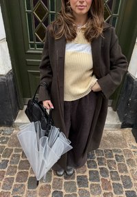 Sac à main noir porté avec un parapluie transparent gris. La tenue comprend un long manteau marron, un pull côtelé crème et un pantalon foncé.