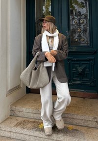 Καφέ βελούδινο σακάκι, λευκό κασκόλ, γκρι φόρμες, μπεζ καπέλο, καφέ γάντια και μια υφασμένη γκρι τσάντα. Στέκεται σε πέτρινα σκαλιά.