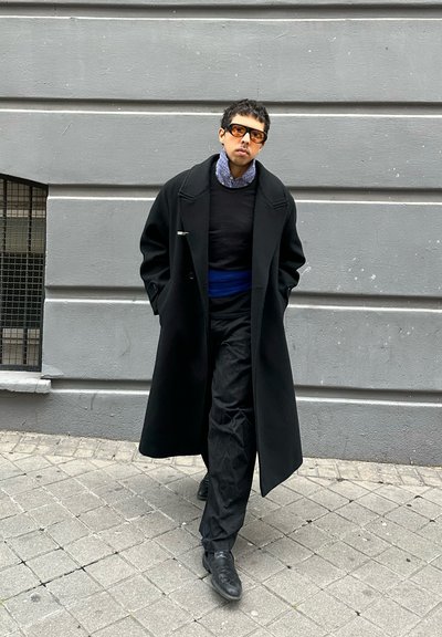 Persona caminando por una acera, vestida con un abrigo negro extragrande, pantalones negros, cinturón azul, camisa con estampado y gafas de sol oscuras, contra un fondo de pared gris.