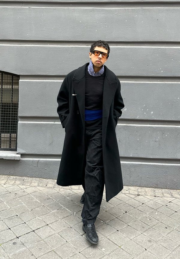 Žmogus vaikšto šaligatviu dėvėdamas per didelę juodą striukę, juodas kelnes, mėlyną diržą, margintą marškinius, tamsius akinius nuo saulės, prieš pilką sienos foną.