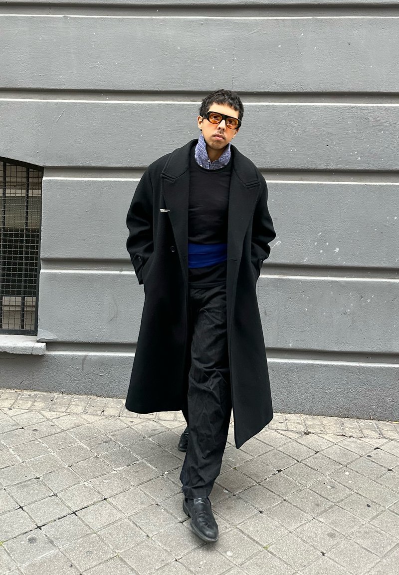 Person, die auf dem Bürgersteig geht, trägt einen übergroßen schwarzen Mantel, schwarze Hose, blauen Gürtel, gemustertes Hemd und dunkle Sonnenbrille, vor grauer Wand.