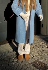 Personne portant un manteau en laine bleu clair, un pantalon crème, des gants beige et des bottes en suède marron, debout sur un pavé près d'une porte.