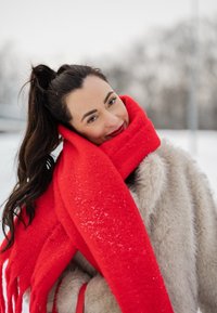 Jeune femme en plein air portant un manteau d'hiver et une écharpe rouge vif, souriante avec un arrière-plan enneigé et flou.