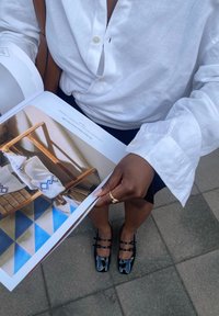 Chemise en lin blanche avec des détails de boutons, chaussures noires à bouts carrés, et un magazine montrant une chaise en bois avec des accents bleus et blancs.