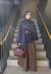 Pelerină bleumarin cu o curea maro, helancă bleumarin și pantaloni largi maro. Geantă de piele maro; scări și balustradă în fundal.